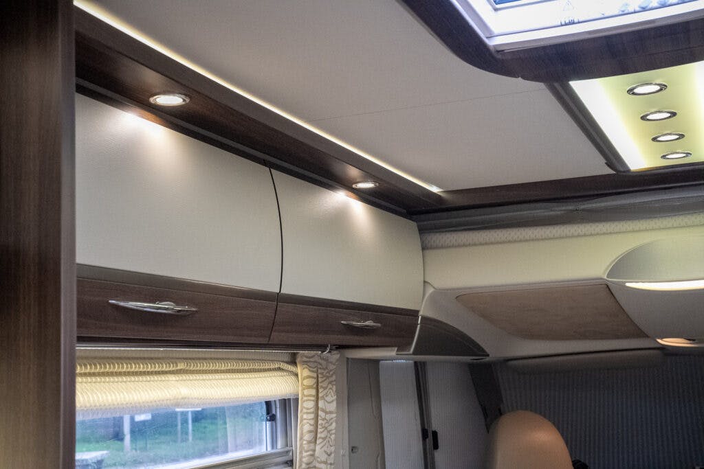 The image shows the ceiling and upper storage cabinets of a 2013 Burstner Elegance 810 G RV interior. The cabinets have a wood finish with curved handles. Recessed lighting and a skylight provide illumination, while a window with a closed, patterned curtain is visible on the left.
