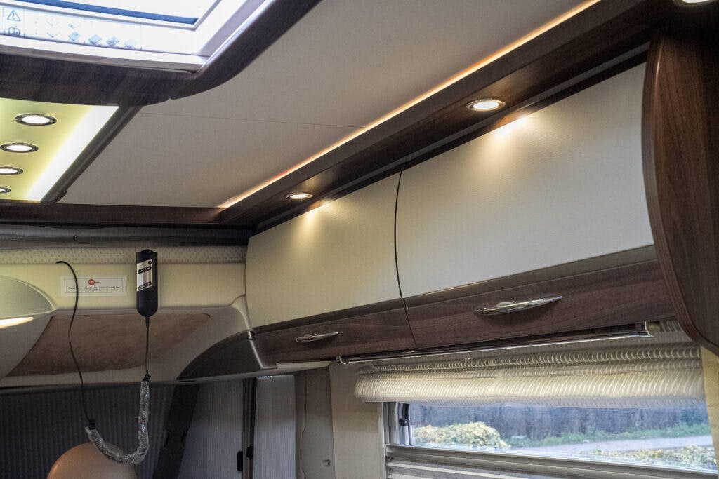 The image showcases the interior ceiling of a 2013 Burstner Elegance 810 G motorhome, featuring overhead storage cabinets with wooden accents and modern ceiling lights. Below the cabinets, a partially visible window with a closed blind is present. The ceiling also has a skylight.
