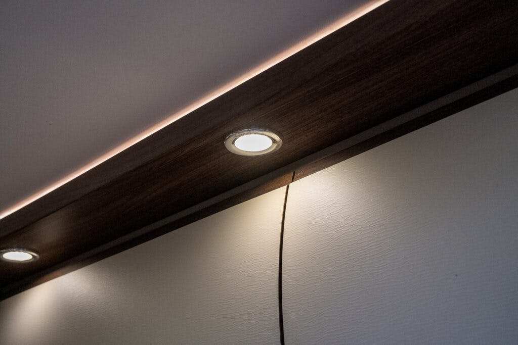A dark wooden ceiling panel with recessed round lights and an adjacent LED strip light adorns the interior of the 2013 Burstner Elegance 810 G. A thin black cable hangs down from the panel against a white textured wall.