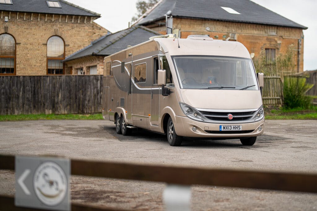 A large beige 2013 Burstner Elegance i810 G motorhome is parked in a lot in front of brick buildings and a wooden fence. The vehicle license plate reads "MK13 HMS." A blurred sign with an arrow is visible in the foreground. The setting appears to be a quiet, possibly rural area.