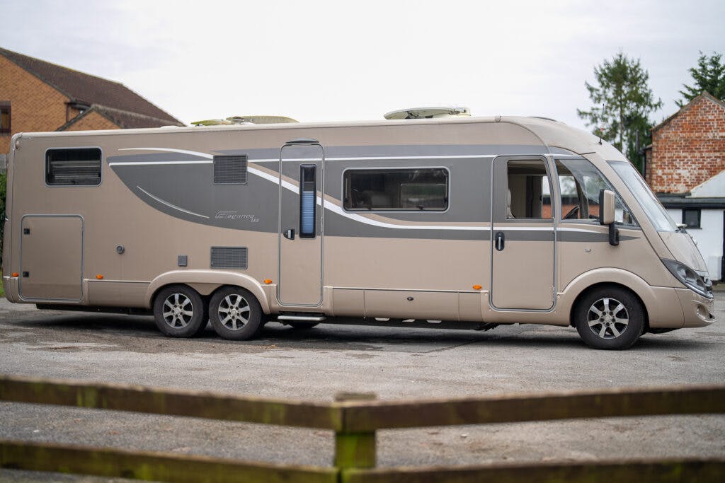A large beige 2013 Burstner Elegance i810 G motorhome with a sleek, modern design is parked in an outdoor area. The vehicle features multiple windows, external storage compartments, and a ladder on the rear. A brick building and a tree can be seen in the background.