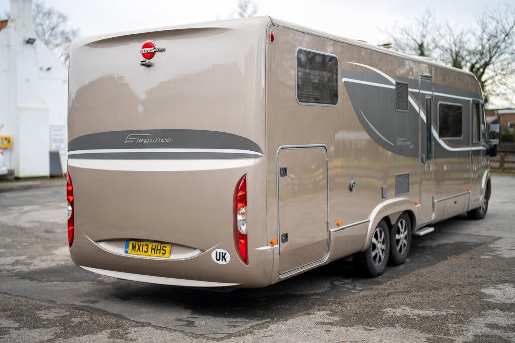 A beige 2013 Burstner Elegance 810 G motorhome is parked on a paved area. The vehicle has two rear wheels on each side and a UK license plate reading "WX13 HHS." It features a sleek design with grey accents, complemented by trees and buildings in the background.