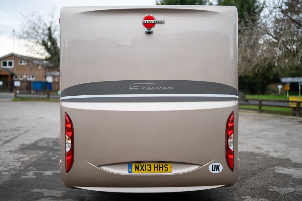 Rear view of a beige 2013 Burstner Elegance 810 G motorhome parked on a street, showcasing the brand logo, "Elegance" lettering, and UK license plate "MX13 HHS". The motorhome features rear lights and a "UK" sticker. Residential houses and trees are visible in the background.