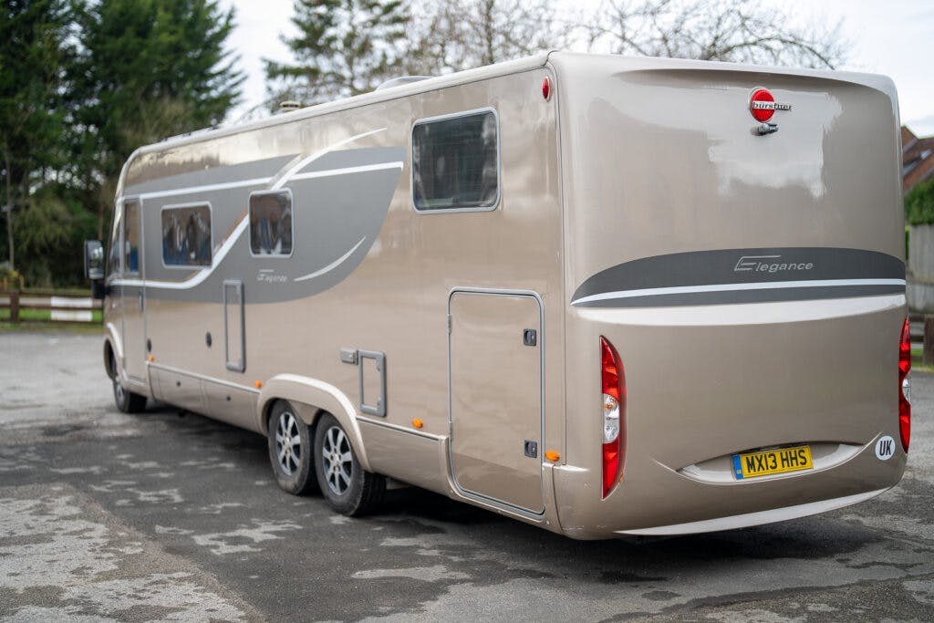 A beige and gray 2013 Burstner Elegance 810 G motorhome with the license plate WX13 HMS is parked on a paved surface. The motorhome features multiple windows, dual rear axles, and an "Elegance" badge on the back. Trees and a wooden fence can be seen in the background.
