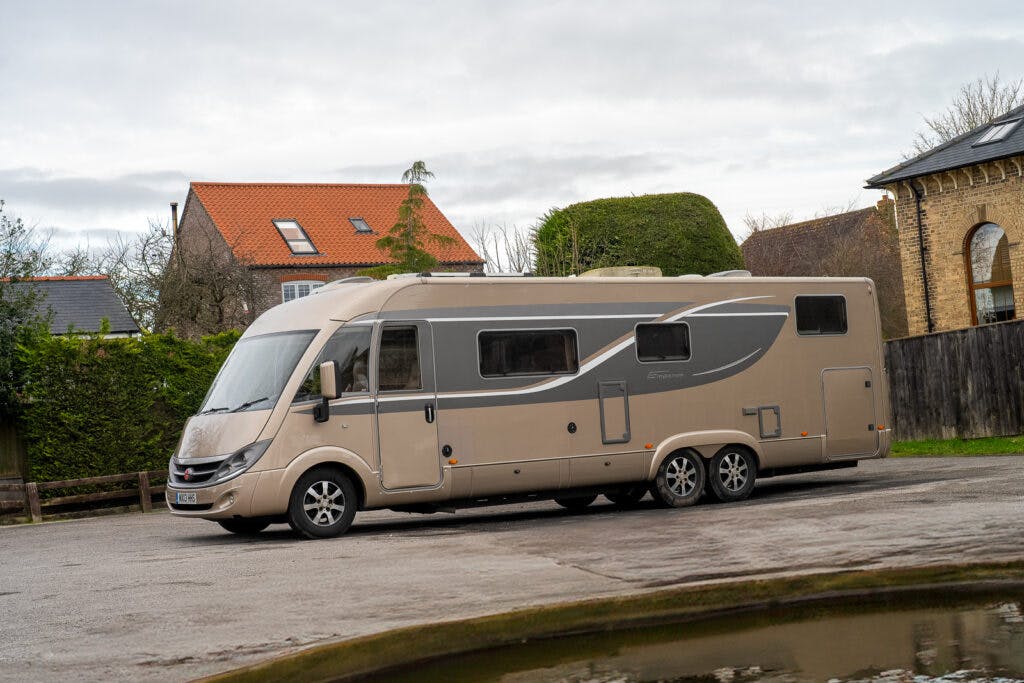 A beige, modern 2013 Burstner Elegance i810 G motorhome with three axles is parked on a paved surface in front of a well-manicured hedgerow and some residential buildings. The sky is overcast.