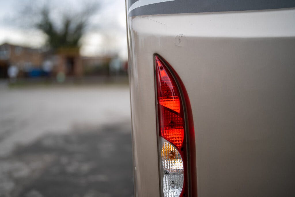 Close-up of the rear taillight on a beige 2013 Burstner Elegance 810 G. The taillight features a red light at the top and white and yellow lights at the bottom. The background is blurred, revealing an outdoor setting with a building and trees.