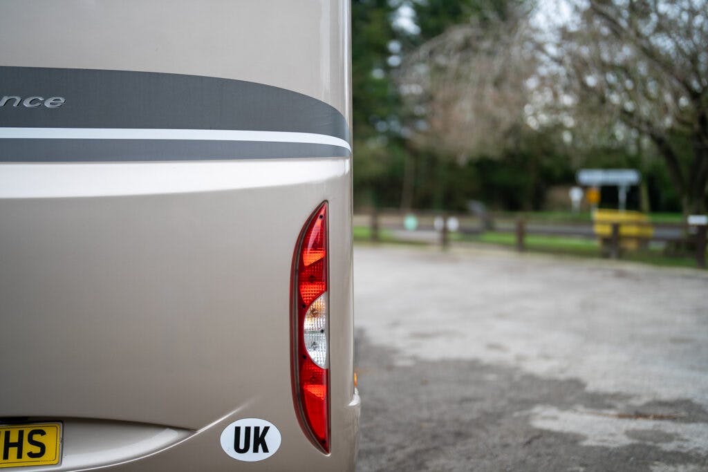 The image showcases the rear portion of a beige 2013 Burstner Elegance 810 G with a UK sticker and a visible taillight. The vehicle is parked on a paved area near a fence, surrounded by trees and greenery. In the distance, there's a partially blurry signpost.