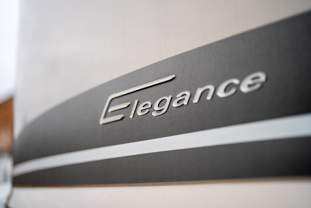 Close-up of a sleek, modern vehicle with the brand name "Elegance" prominently displayed in metallic letters on a black and white background. The focus is on the brand name of the 2013 Burstner Elegance 810 G, highlighting its stylish and sophisticated design.