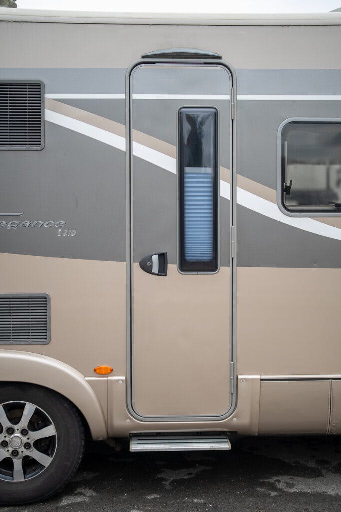 A close-up view of the side door of a 2013 Burstner Elegance 810 G RV. The door features a grey and beige color scheme, with a vertical window in the center and a small step below. Part of a wheel and side window are visible.