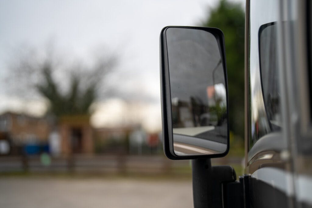 A close-up of a black side mirror attached to a 2013 Burstner Elegance 810 G. The background is blurred, showing an outdoor scene with some buildings, trees, and a fence. The image is taken from the side of the vehicle, focusing on the mirror's reflective surface.
