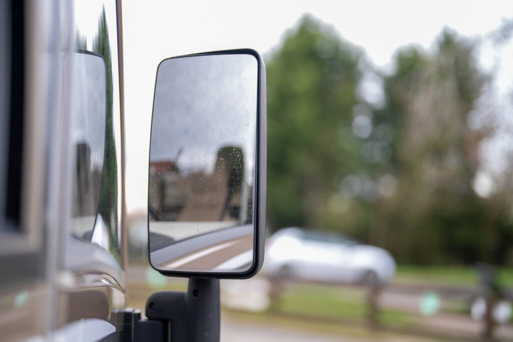 Close-up of a vehicle's side mirror showing a slightly blurred, rainy reflection. In the mirror and the background, there are trees and another vehicle, suggesting a serene outdoor environment. The 2013 Burstner Elegance 810 G adds an air of sophistication to this picturesque scene.