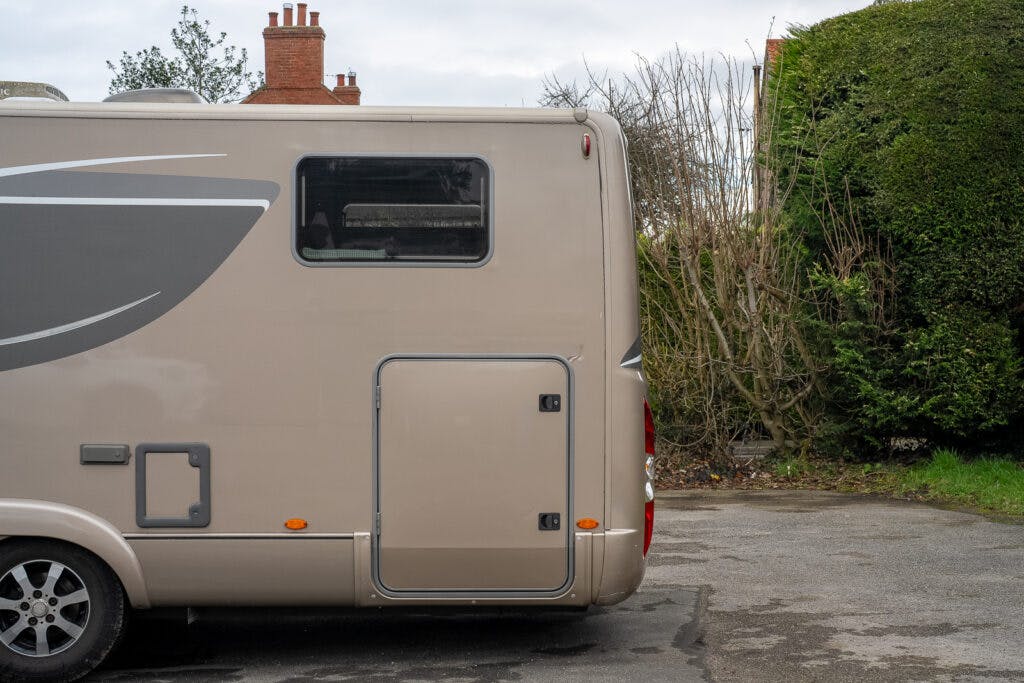 Rear and side view of a beige 2013 Burstner Elegance i810 G motorhome parked on a driveway. The motorhome has a small window, storage compartments, and red taillights. In the background, there are brick chimneys and a large hedge. The driveway surface appears worn and slightly uneven.
