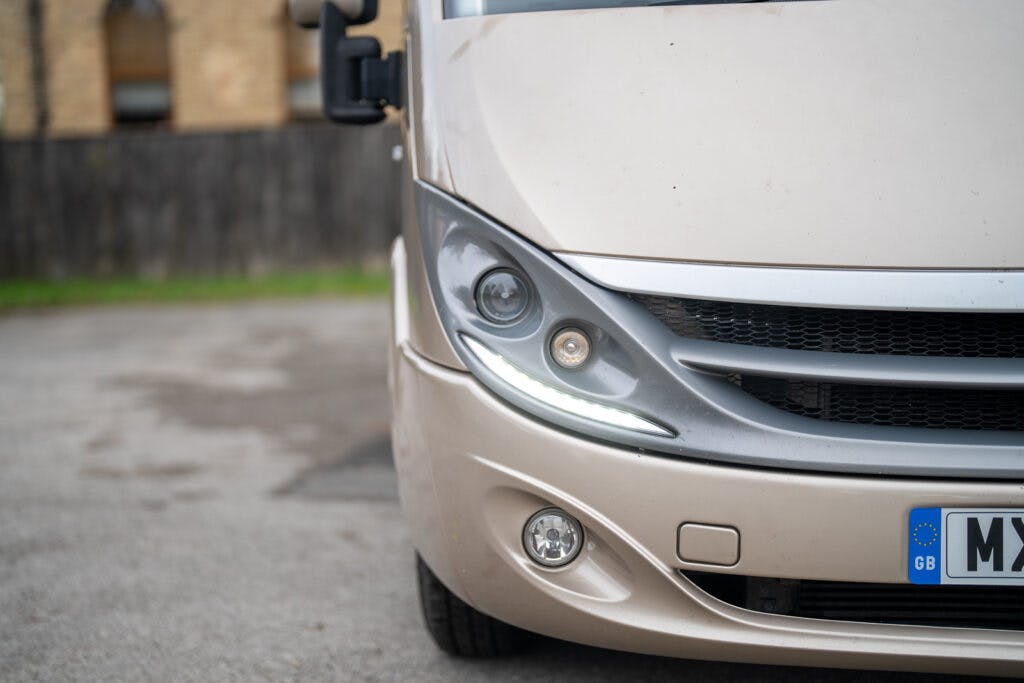 The image shows a close-up of the front left side of a beige vehicle, specifically the 2013 Burstner Elegance 810 G, focusing on the headlight and part of the grille. The vehicle has a European license plate with a 'GB' designation partially visible on the right side. A blurred brick building is in the background.