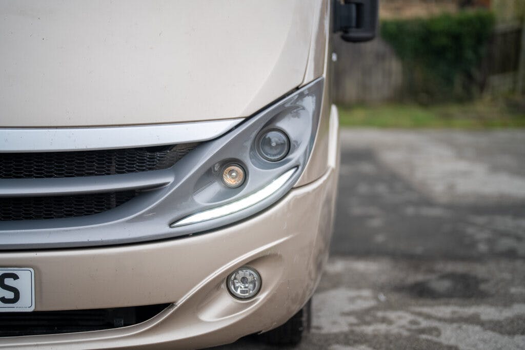 A close-up of the front right side of a beige 2013 Burstner Elegance 810 G RV, showing its headlight, fog light, and part of the grille. The surrounding area seems to be an outdoor setting with a paved surface and a blurred background with greenery.