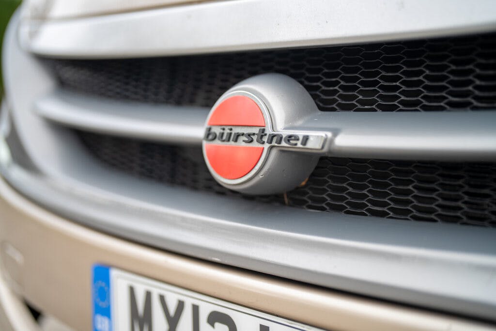 Close-up of a Bürstner vehicle badge on the front grille of a 2013 Burstner Elegance 810 G. The logo consists of a red circle with the brand name "Bürstner" in silver and black lettering across the middle. Part of a European license plate is visible below the grille.