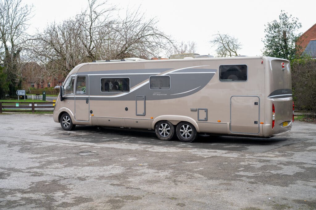 A beige and gray 2013 Burstner Elegance 810 G recreational vehicle (RV) is parked in an empty lot. The RV features multiple windows, sleek design elements, and dual rear wheels. Leafless trees and a building with a red roof are visible in the background.