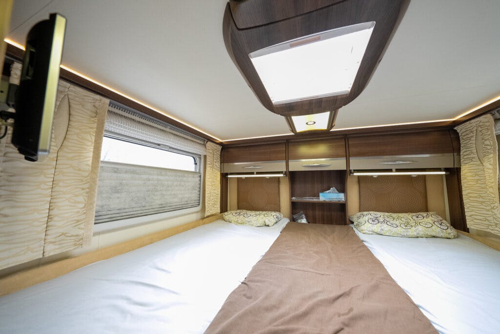 Interior view of the 2013 Burstner Elegance 810 G camper van bedroom with two single beds separated by a narrow aisle. Wooden cabinets and shelves are above the beds, which have pillows and a brown runner. A window with patterned curtains is on the left side, with a television mounted next to it.