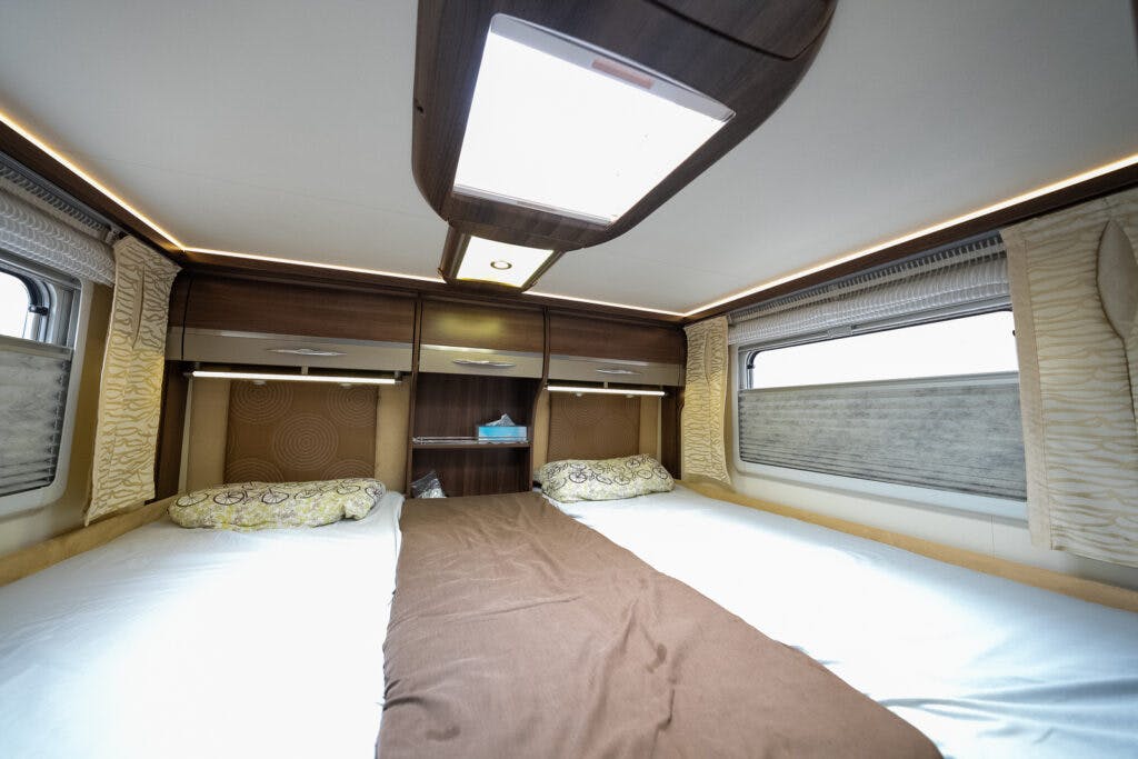Interior of the 2013 Burstner Elegance 810 G features a double bed arrangement with two twin mattresses, each with pillows and a shared brown blanket in the middle. The room boasts wooden cabinets, windows on both sides, and a skylight above.