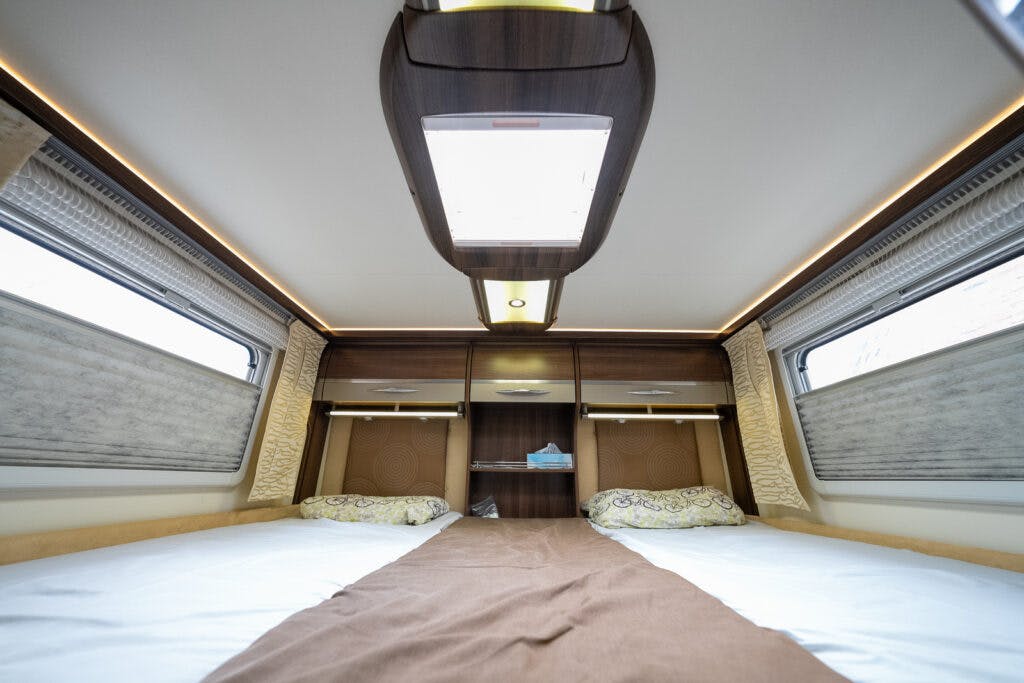 The image shows the interior of a 2013 Burstner Elegance 810 G bedroom with two beds, a shared brown blanket, and patterned pillows. Wooden cabinets and shelves with accent lighting line the back wall. Two windows with shades are on either side.