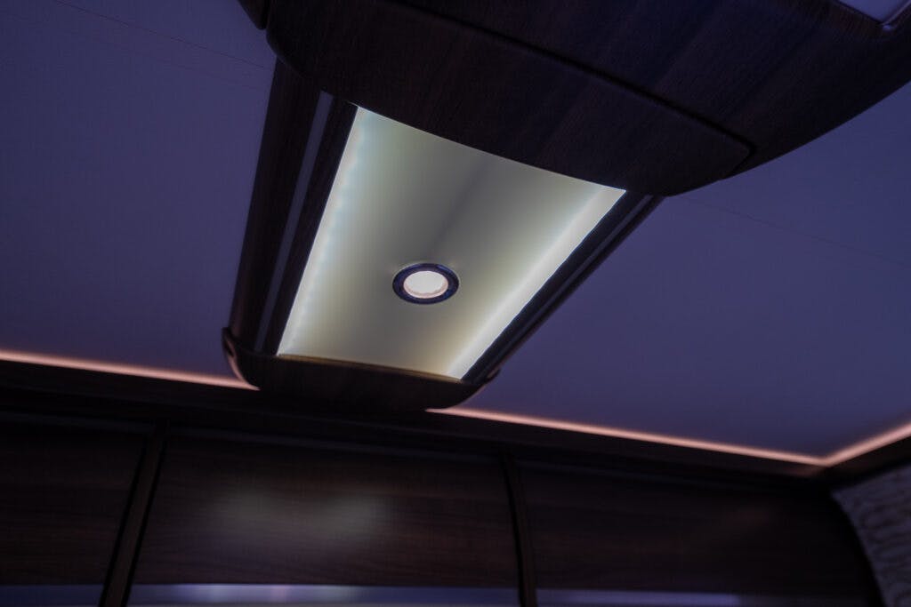 A modern ceiling light fixture with a sleek, rectangular design and dark wooden accents, reminiscent of the luxury seen in a 2013 Burstner Elegance 810 G. The fixture features a central circular light surrounded by a soft, glowing perimeter, providing ambient illumination in the room.