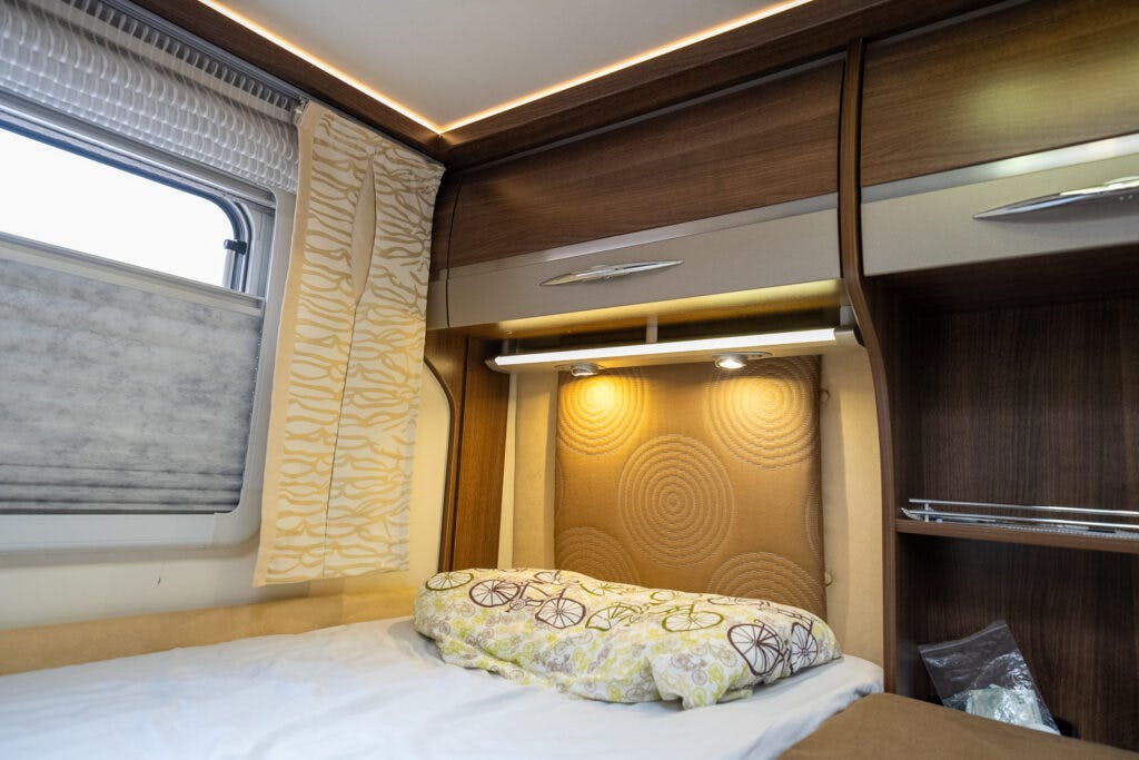 The image shows the interior of a 2013 Burstner Elegance 810 G camper van bedroom with a neatly made bed. The bed has a white sheet and a decorative pillow with floral patterns. The walls are lined with wooden cabinets, and there is a window with patterned curtains. Two overhead lights are on.
