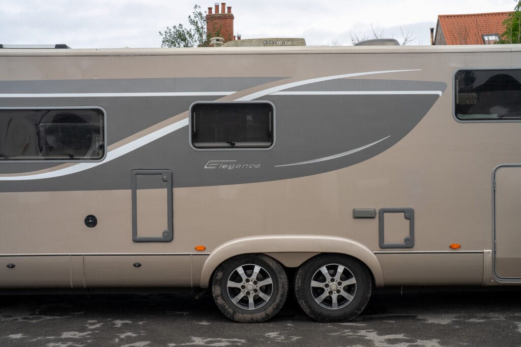 A beige 2013 Burstner Elegance i810 G motorhome with gray and white accents parked on a paved surface. The vehicle features two rear wheels, windows, and various utility access panels. The word "Elegance" is written on the side. Background includes part of a roof and sky.