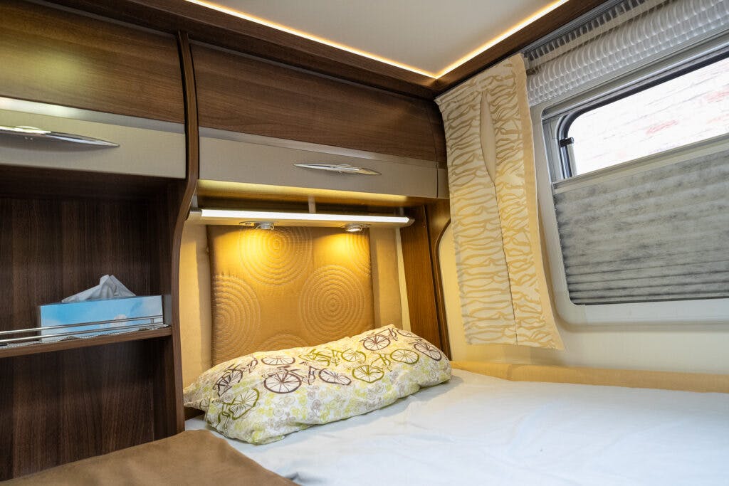 The image captures the interior of a 2013 Burstner Elegance 810 G camper van bedroom. It features a single bed with a bicycle-themed pillow, an overhead shelf with lighting, a box of tissues on a shelf, and a window with patterned curtains and blinds. The design boasts wood and beige tones.