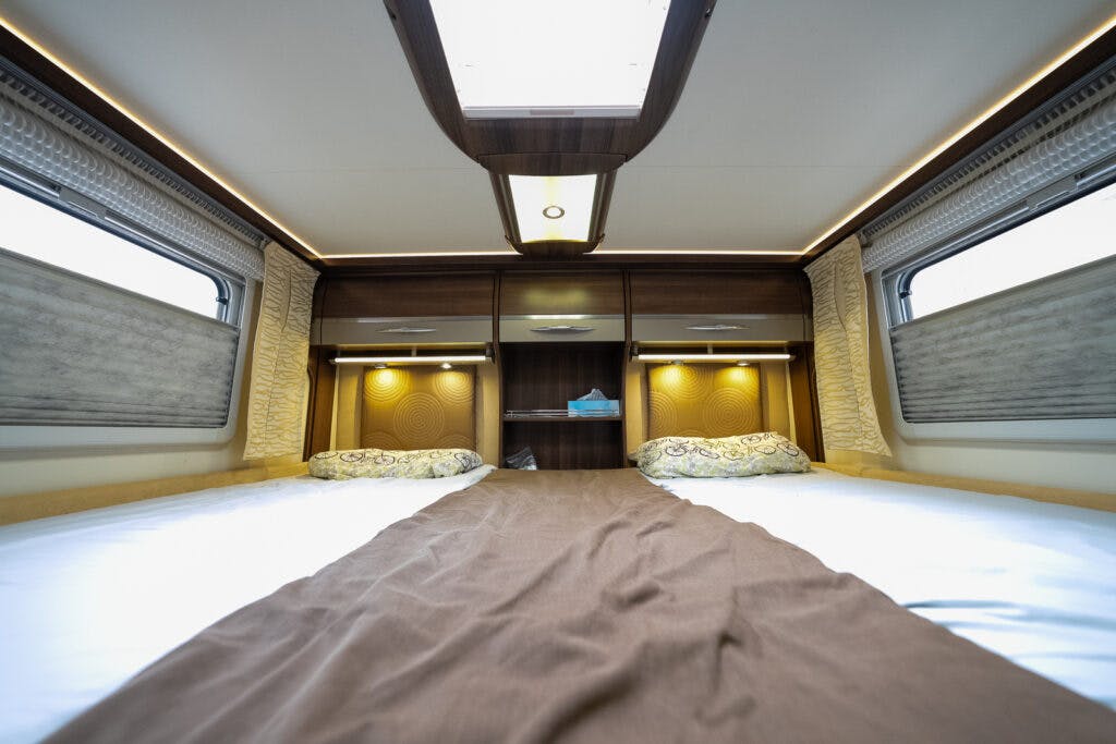 Interior of the 2013 Burstner Elegance 810 G camper van bedroom with two single beds separated by a center aisle. The beds have brown and white bedding, decorative pillows, and overhead storage compartments. Two windows let in natural light, creating a bright and cozy atmosphere.