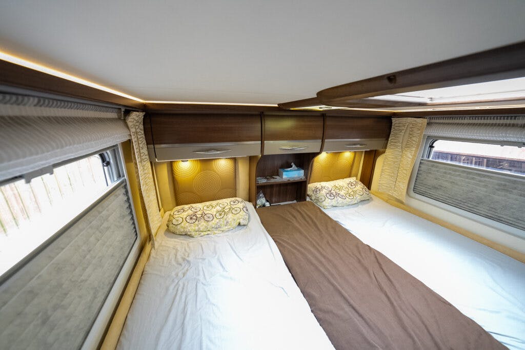 The image shows the interior of a 2013 Burstner Elegance 810 G motorhome bedroom with two single beds separated by a narrow brown runner. The beds have patterned pillows and there are overhead cabinets with built-in lights. A small shelf with a tissue box is positioned between the beds.