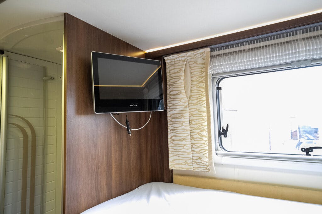 A small television is mounted on a wooden wall inside the 2013 Burstner Elegance 810 G RV. Next to the television, a window with patterned curtains is partially drawn. Below the TV, there is a bed with a white blanket, and the adjacent bathroom door is partially visible.