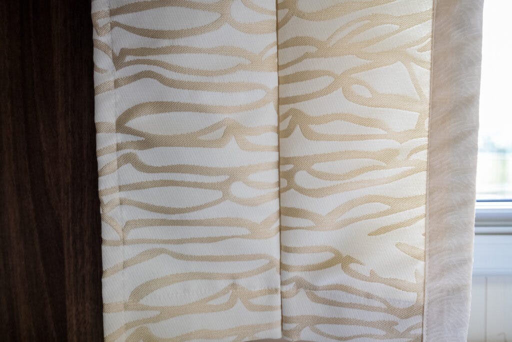 A close-up view of a light-colored curtain with a wavy, abstract pattern in beige and white inside a 2013 Burstner Elegance 810 G. The curtain is partly drawn, revealing a white window frame on the right side and a wooden surface on the left. The scene is lit by natural light.