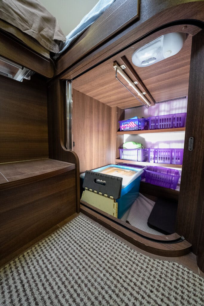 A small enclosed storage space with wooden walls featuring organized shelves. The shelves hold plastic baskets in purple and white, and a blue storage container. The carpeted floor mirrors the cozy feel of a 2013 Burstner Elegance 810 G, lit by a small ceiling light and a strip light above the shelves.