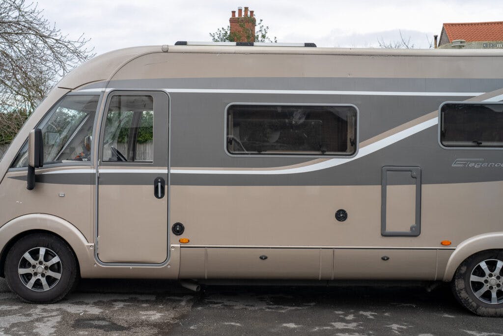 Side view of a beige and gray 2013 Burstner Elegance i810 G motorhome parked on a road. The vehicle has large windows and a door on the left side, with a visible tire and part of a house's roof in the background. The motorhome's surface shows some signs of dirt and wear.