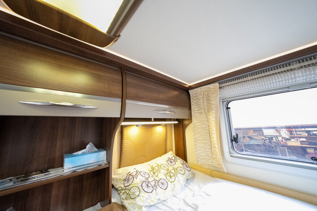 The interior of the 2013 Burstner Elegance 810 G features a built-in wooden cabinet and lighted shelving above a cozy bed. Beside the bed is a window with a curtain, while the bedding includes a patterned pillow and a light blanket. Tissues and other small items rest neatly on the shelf.
