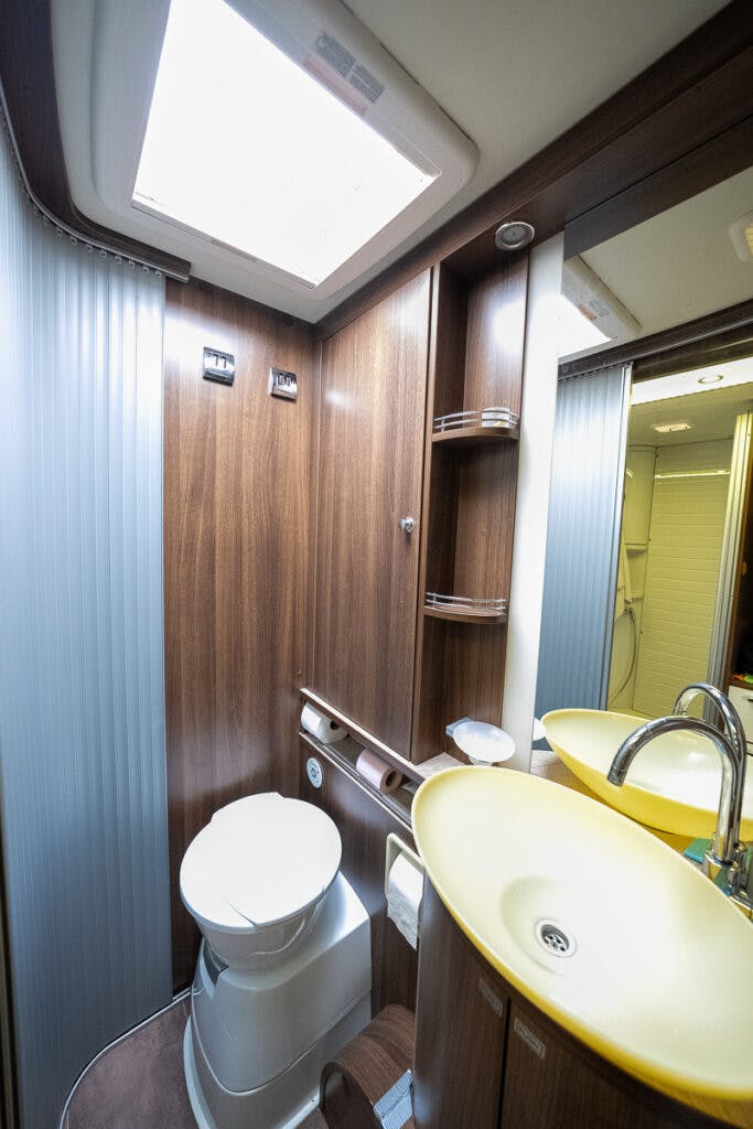 A compact bathroom in the 2013 Burstner Elegance 810 G features a wooden interior, flush toilet, and yellow sink with a chrome faucet. Above the sink, shelves are integrated into the wall. The ceiling has a skylight, and a mirrored cabinet is present on the right.