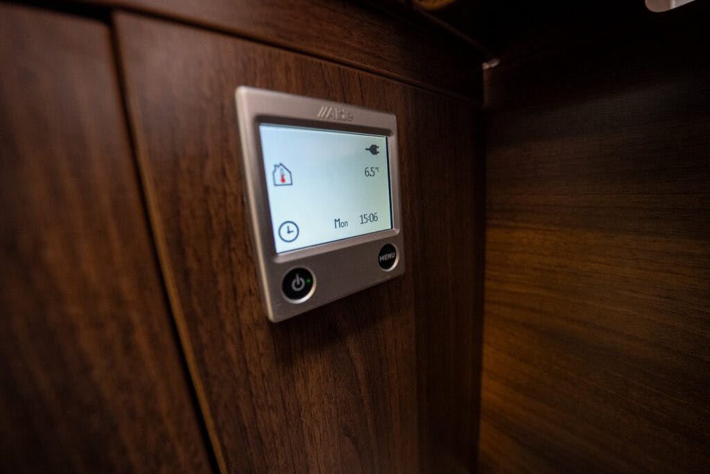 A digital control panel mounted on a dark wood surface in the 2013 Burstner Elegance 810 G displays a temperature of 65.7 degrees Celsius. The panel shows icons for home and a timer reading 150:16. Below the screen are power and menu buttons, with the panel branded with "Alde.