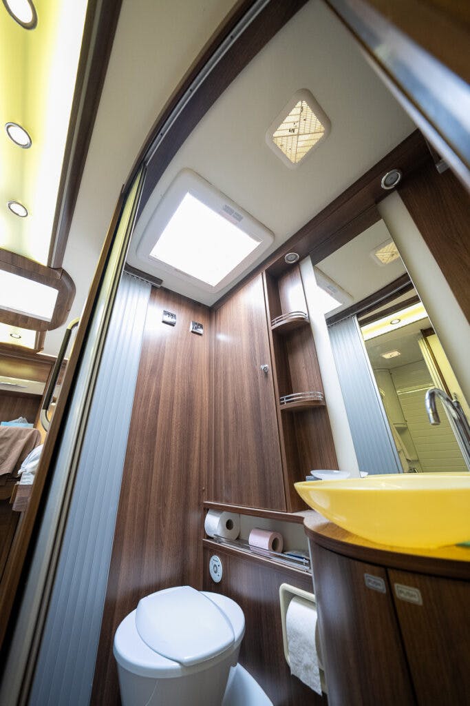 This image shows a compact, modern bathroom with wooden paneling in the 2013 Burstner Elegance 810 G. There is a toilet, a small sink with a yellow basin, two shelves, and a mirror. A window on the ceiling allows natural light to enter the space, which is reflected in other mirrored surfaces.