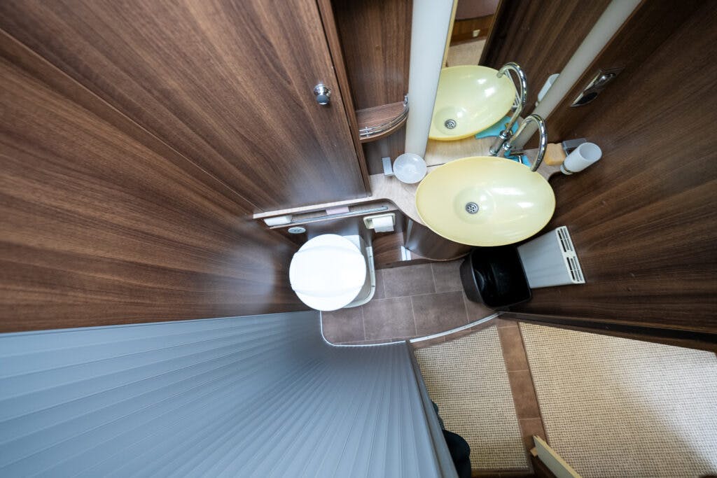 A bird's-eye view of the compact bathroom in the 2013 Burstner Elegance 810 G. The space features a toilet, a small yellow sink, and wooden cabinetry. The floor is tiled, and a portion of the adjacent room with carpeted flooring is visible at the bottom right.