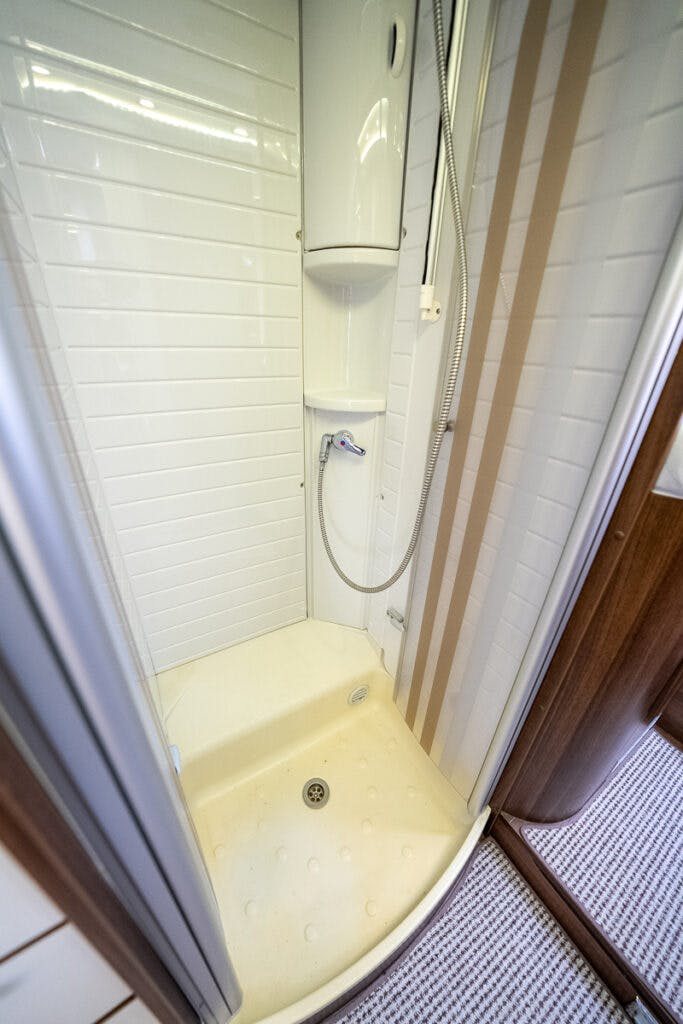 The 2013 Burstner Elegance 810 G features a compact, white-tiled shower with a sliding glass door, a small shelf, and a handheld showerhead attached to a flexible hose. The shower has a beige-colored base with a drain in the center and is situated in an area with wooden paneling and carpeted flooring.