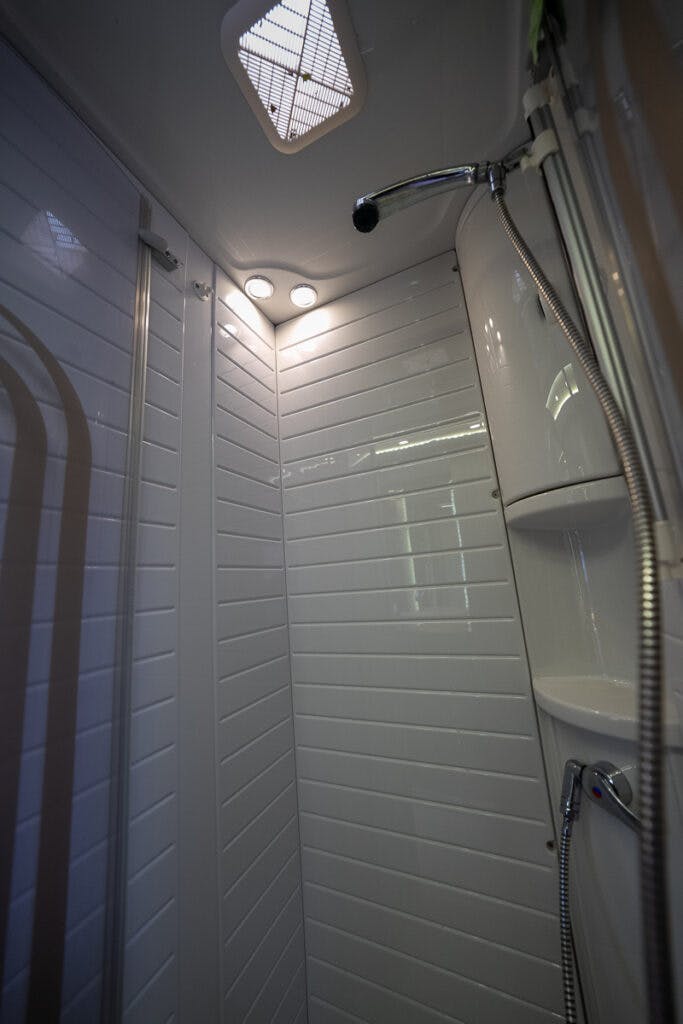 A compact shower stall in the 2013 Burstner Elegance 810 G features white paneled walls, a chrome showerhead, and a detachable handheld showerhead. The stall includes built-in corner shelves, overhead lighting, and a small ventilation window on the ceiling.