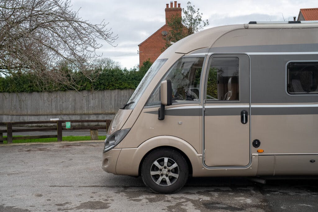 A parked beige and grey 2013 Burstner Elegance i810 G motorhome is positioned sideways on a paved area. Behind the vehicle is a wooden fence with trees and bushes visible above it. The background includes a building with a red brick exterior and a chimney.