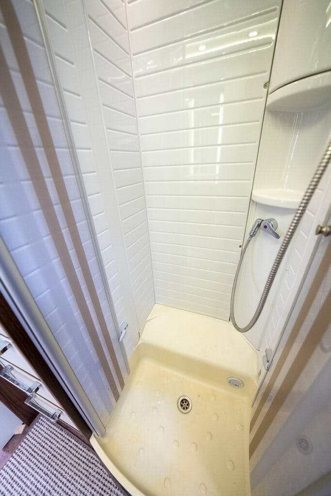 A compact, white-tiled shower with a sliding glass door in the 2013 Burstner Elegance 810 G. The shower features a handheld shower head attached to a flexible hose, mounted on the wall. There are built-in shelves in the corner, and the shower floor has a central drain.