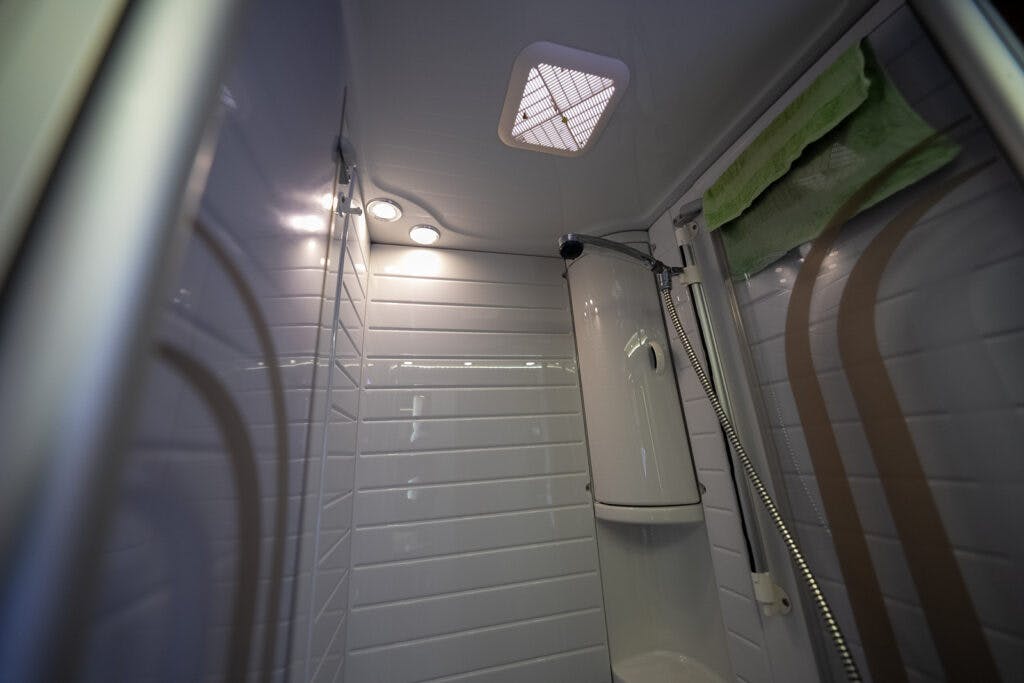 A small, modern shower stall with white tiled walls and a ceiling vent, reminiscent of the sleek design found in a 2013 Burstner Elegance 810 G. The shower features a vertical water heater, a flexible shower hose, and a green towel hanging on a bar. Recessed lighting provides illumination inside the stall.