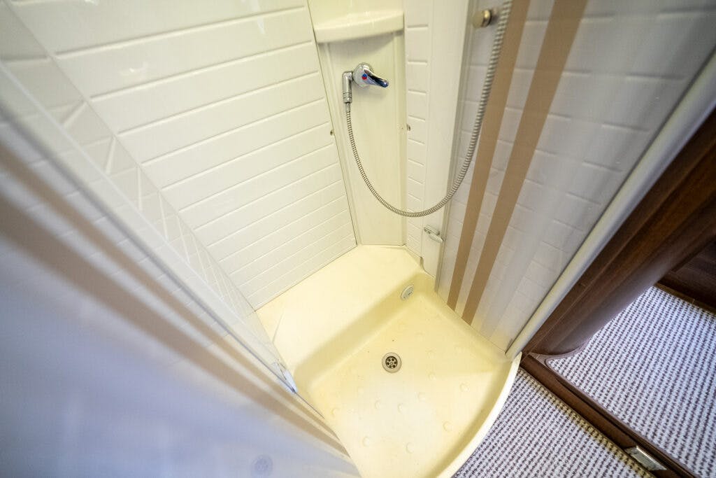 A compact shower stall in the 2013 Burstner Elegance 810 G features white tiles on the walls. The showerhead is attached to a flexible hose mounted on a wall bracket. The floor of the stall is slightly sloped towards the drain, and the area is enclosed by glass panels.