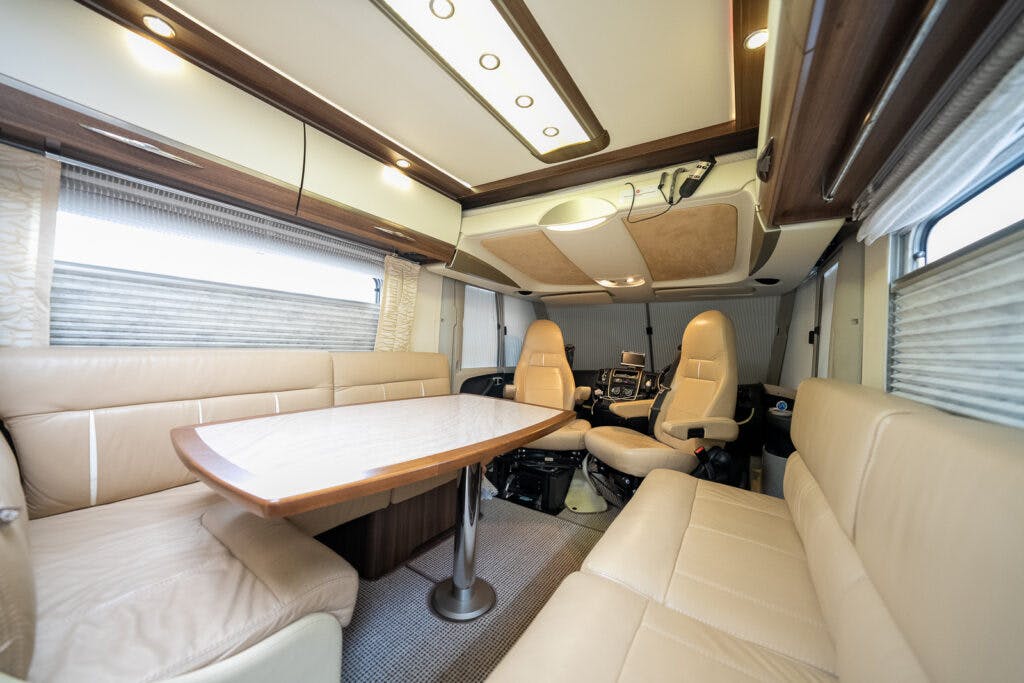 The 2013 Burstner Elegance 810 G boasts a spacious RV interior, featuring beige leather seating around a wooden table, with additional seats at the front near the driver's area. The windows have blinds, and the ceiling has recessed lighting and storage compartments.