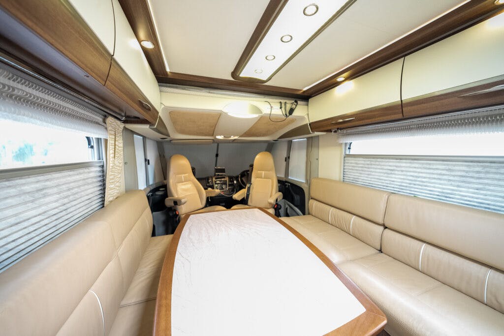 The interior of the 2013 Burstner Elegance 810 G RV features a dining area with a long table surrounded by beige leather seating. The driver and passenger seats can be seen at the far end, with large windows along both sides admitting light. The ceiling has built-in lighting fixtures.