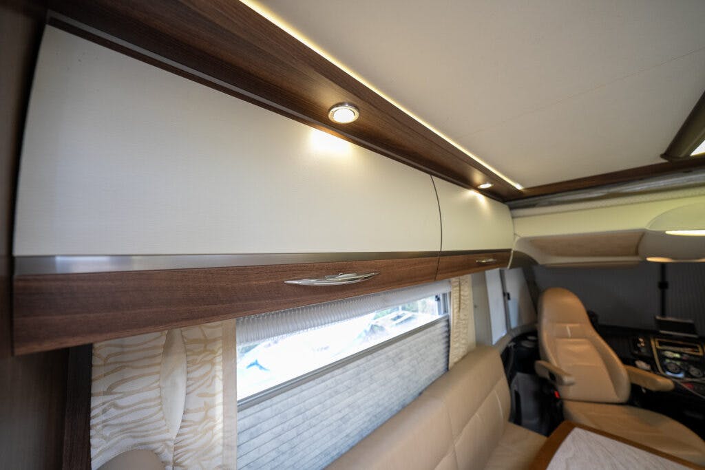 Interior of a 2013 Burstner Elegance 810 G motorhome featuring a white and woodgrain upper storage cabinet with modern lighting fixtures. A cream-colored cushioned seat is seen below, along with a large window with a patterned curtain and another seat partially visible on the right.