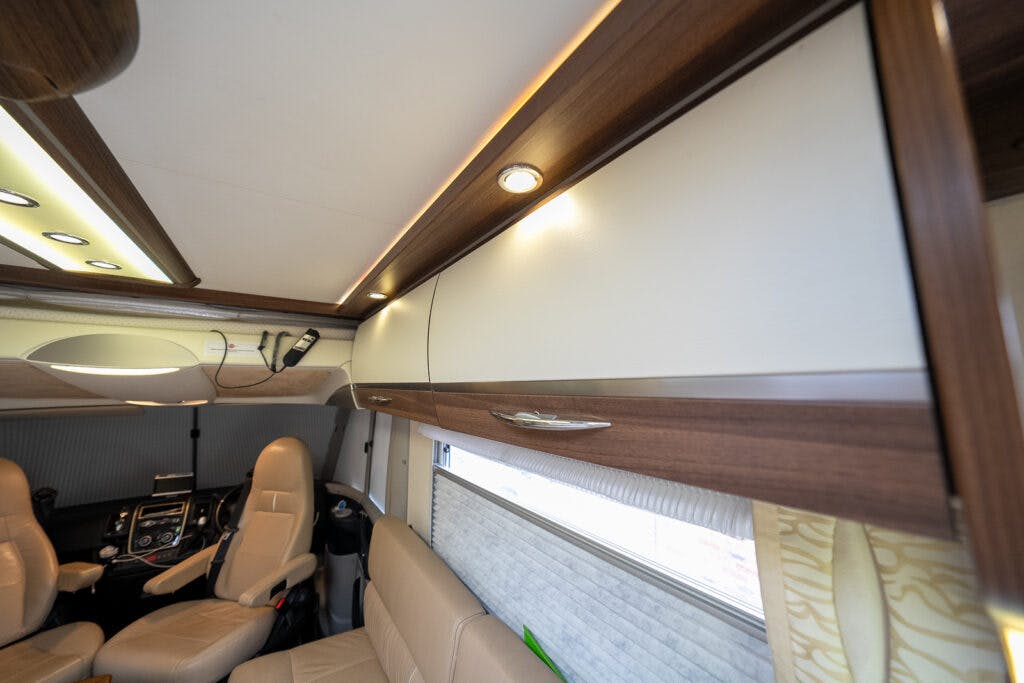 The interior view of the 2013 Burstner Elegance 810 G motorhome features beige leather seats, overhead storage cabinets with wood trim, and a cockpit area with various driving controls. There are ceiling lights and a window with a pull-down shade.