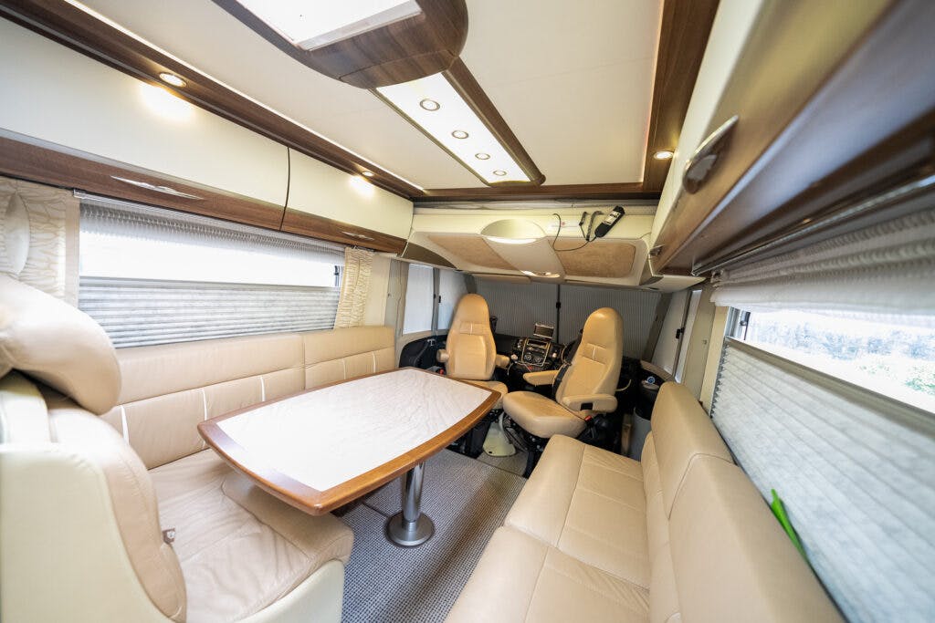 The interior of the 2013 Burstner Elegance 810 G RV boasts a modern living space with tan leather seating. It features a U-shaped sofa around a wooden table, two swivel chairs, overhead cabinets, and windows with shades. The space is well-lit with ceiling lights.