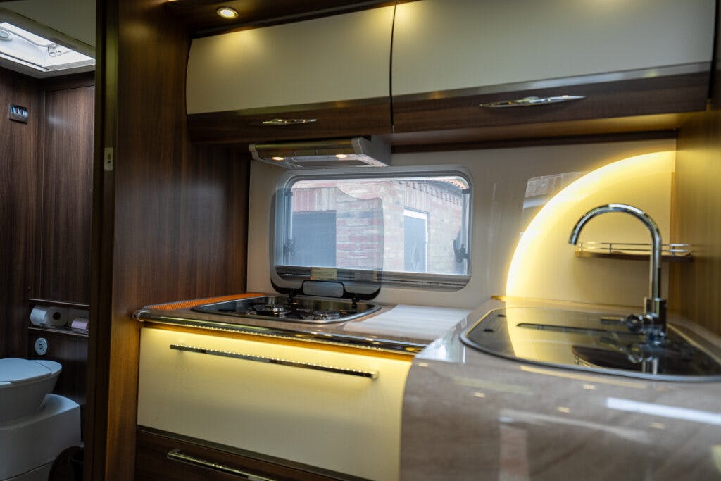 This image shows the interior of a 2013 Burstner Elegance 810 G kitchen. It features a sink with a metallic faucet, a gas stove with two burners, and wooden cabinets with built-in lighting. A window provides a view of an outdoor brick wall. There is also a small toilet area visible.